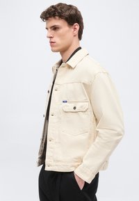 Νέος άνδρας με καστανά σγουρά μαλλιά, που φοράει εκρού τζιν μπουφάν και μαύρο παντελόνι, στέκεται προφίλ με τα χέρια στις τσέπες.