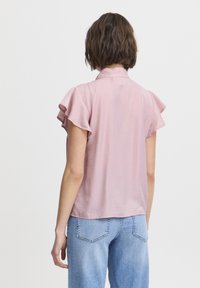 Blusa a maniche corte di un delicato rosa chiaro con maniche a sbuffo, colletto e una texture liscia. Abbinata a jeans azzurri chiari.