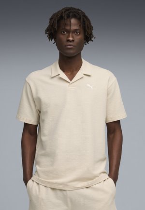 Młody mężczyzna z krótkimi dredami w teksturowanej kremowej koszulce polo z krótkim rękawem i pasujących spodniach, z rękami w kieszeniach.