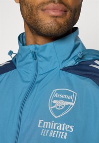 Blauwe ritsjack met een hoge kraag, voorzien van het Arsenal-logo en de tekst "Emirates Fly Better" in het wit. Glad materiaal met contrasterende marineblauwe schouderaccenten.