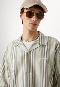 Chemise à rayures vertes et beiges à manches courtes avec col, fermeture à boutons et logo brodé. Modèle portant des lunettes de soleil et une casquette.