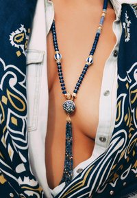 Collier perlé bleu et blanc avec un pendentif rond à motif et un long gland. Comprend des accents dorés et des tailles de perles variées, reposant sur un tissu.