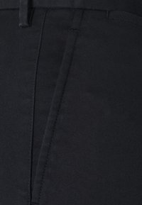 Detailní záběr na černé kalhoty, ukazující přední zip, poutka na pásek a precizní šití na látce.