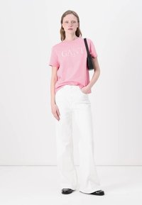 Ljusrosa bomulls-T-shirt med vit logotyp, i kombination med höga vita vida jeans och svarta sandaler med öppen tå, modellen håller en liten svart väska.