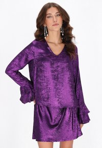 Violettes, metallisches Kleid mit langen Ärmeln und Rüschenakzenten. Es hat einen tiefen V-Ausschnitt und eine lockere Passform, gefertigt aus einem glatten, glänzenden Stoff.