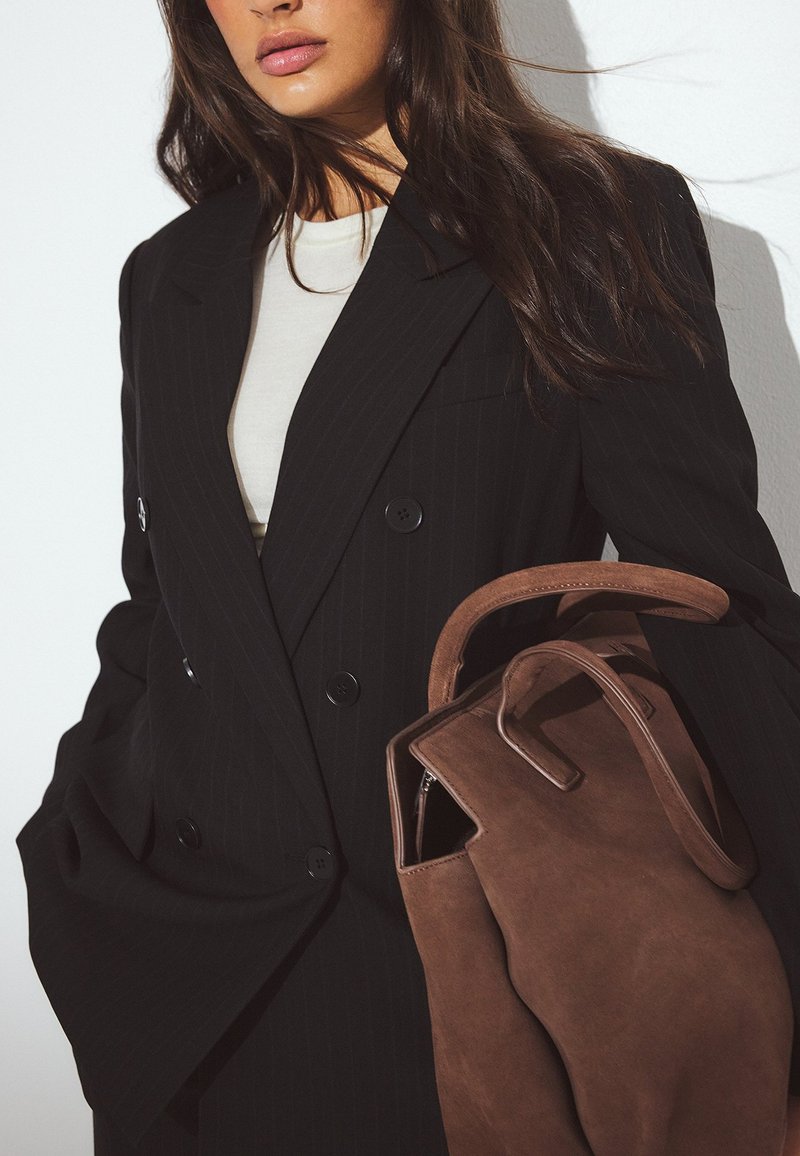 Kobieta w czarnym, dwurzędowym żakiecie w drobną prążek, trzymająca dużą brązową zamszową torebkę z zaokrąglonymi uchwytami.