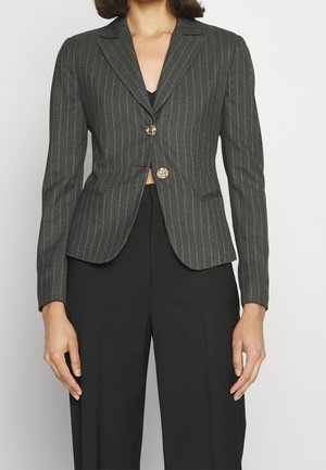 Kobieta ubrana w dopasowaną ciemnoszarą marynarkę w cienkie prążki z dwoma złotymi guzikami, noszoną na czarnych spodniach z wysokim stanem.