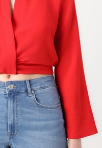 Blouse rouge courte à manches longues, en tissu texturé, associée à un jean denim bleu taille haute avec un design classique de bouton et de poches.