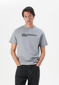 Gai pelēks kokvilnas T-krekls ar īsām piedurknēm un apaļu kakla izgriezumu. Priekšpusē ir uzdrukāts raupjš melns teksts "KARL LAGERFELD JEANS". Saskaņots ar melniem džinsiem.