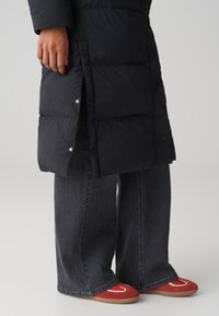 Doudoune noire avec boutons-pression, associée à un jean en denim noir à jambes larges et des baskets rouges avec des accents blancs. Textures lisses visibles.
