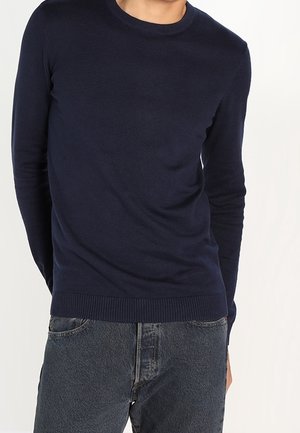 Pull en tricot bleu marine avec un col rond, des manches longues et des poignets côtelés. Porté avec un jean en denim foncé, présentant une coupe ajustée.