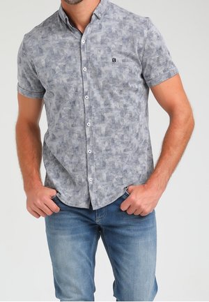Homme portant une chemise à manches courtes à boutons avec un motif abstrait gris et un jean bleu, les mains dans les poches avant.