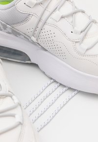 Bílá sportovní podrážka boty stoupá na tři paralelní bílé tkaničky na čistém, světlém šedém povrchu.