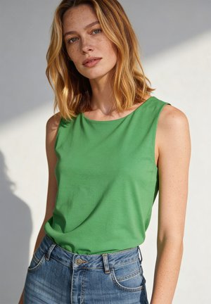 Woman with shoulder-length blonde hair wearing a green sleeveless top and blue jeans against a light background.