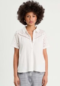 Chemise blanche à manches courtes, avec un tissu texturé, des détails en dentelle à œillets sur les épaules et une coupe décontractée.