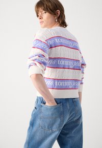 Crème sweater met een getextureerd patroon, met banden van paarse en rode strepen met de tekst "tommy jeans". Gepaard met blauwe denim jeans.