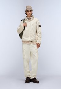 Geacă din polar crem, cu fermoar și buzunare laterale; pantaloni cargo în off-white, cu buzunare laterale cargo; pantofi maronii; rucsac verde.
