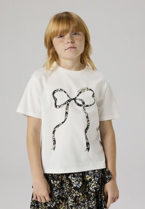 Young red-haired child with freckles wearing a white shirt with black floral bow design and a dark floral skirt on neutral background.