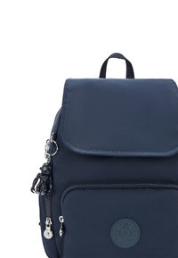 Marinblå tygryggsäck med klaffstängning, framfickor med dragkedja, gummiloggopatch och en dekorativ svart charm fäst vid dragkedjan.