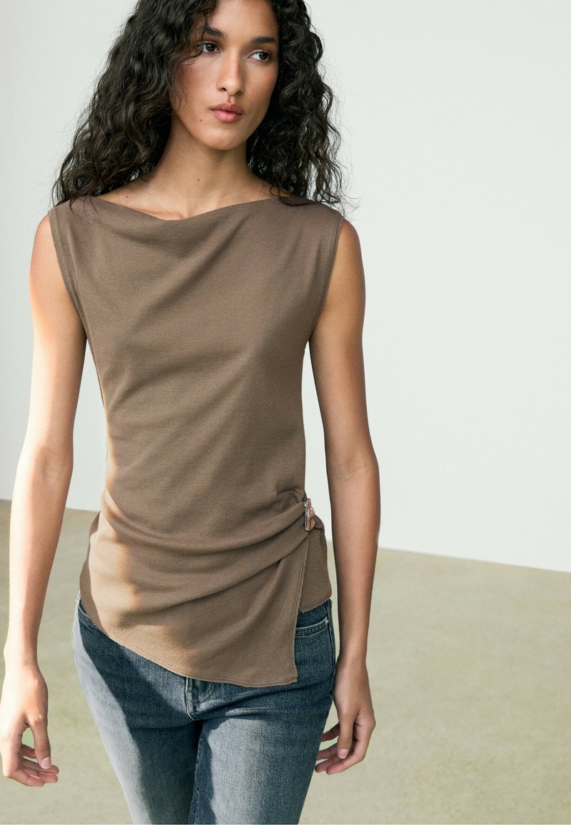 Ung kvinde med krøllet hår iført en ærmeløs brun top med asymmetrisk skæring og blå jeans mod en ensfarvet baggrund.