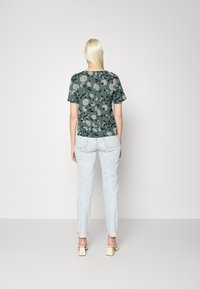 Top verde de mangas curtas com padrão floral sobre um fundo claro, combinado com jeans de ganga clara e sandálias de salto bloco, visto de trás.