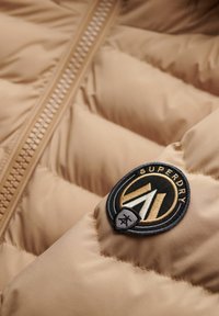 Primer plano de una chaqueta de plumas beige con textura acolchada, que cuenta con un parche circular del logo de Superdry en negro, dorado y blanco cerca de la cremallera.