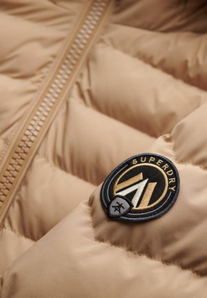 Beige pufferjack met een zichtbare rits, doorgestikt patroon en een ronde zwarte en gouden logopatch met de tekst "SUPERDRY".