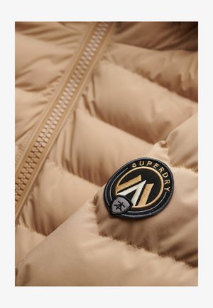 Beige pufferjack met een zichtbare rits, doorgestikt patroon en een ronde zwarte en gouden logopatch met de tekst "SUPERDRY".