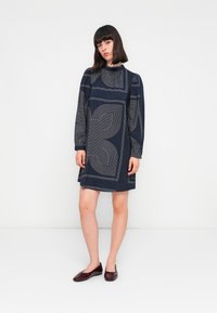 Abito blu navy a tubino con maniche lunghe, caratterizzato da motivi geometrici bianchi e un collo alto. Abbinato a ballerine rosso scuro.