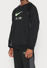 Černá mikina s kulatým výstřihem, která má velké zelené logo Nike a text "AIR". Obsahuje tmavě šedé postranní klapušky a žebrované manžety.