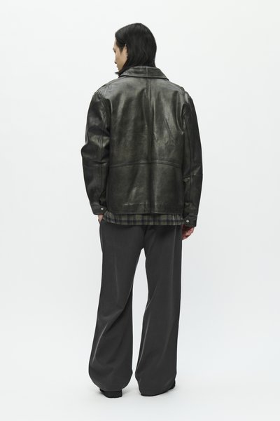 Person with shoulder-length dark hair wearing a black leather jacket, plaid shirt, and loose dark trousers standing facing away.