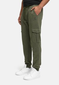 Man in olijfgroene cargo joggers met zijzakken en witte sneakers, staand met één hand in de zak op een witte achtergrond.