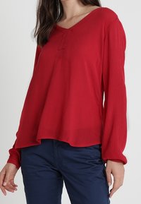 Blusa rossa a maniche lunghe con scollo a V e tessuto strutturato. Presenta un dettaglio con bottone sullo scollo e una vestibilità morbida, abbinata a pantaloni blu navy.