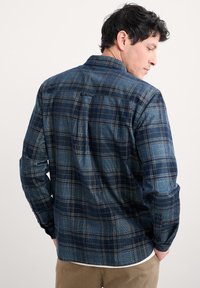 Man in een blauw en zwart geruit overhemd met lange mouwen en een beige broek, met zijn rug naar de camera, met zijn handen in zijn zakken tegen een effen achtergrond.