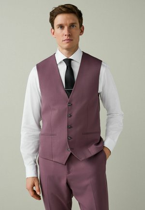 Man wearing mauve suit vest and pants, white shirt, and black tie, standing with one hand in pocket against plain gray background.