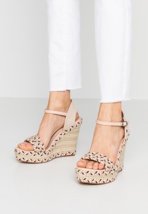 Sandalias de cuña espadrille tejidas con tiras en el tobillo, usadas con pantalones blancos recortados sobre un suelo gris claro.
