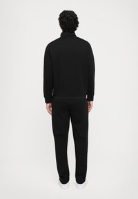 Sweat-shirt et pantalon noirs en tissu doux ; poignets et ourlet ajustés ; design uni sans motifs ; modèle de dos, face à la caméra.