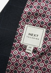 Sötétkék, struktúrált anyag, bordeaux és szürke mintázott béléssel. Fehér márkacímke találhat�ó rajta, "NEXT TAILORING" felirattal és "40R" mérettel.