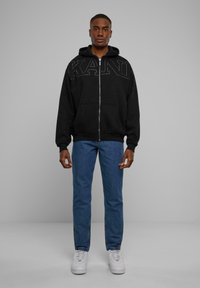 Black zip-up hoodie with studded lettering, paired with blue jeans and white sneakers. The hoodie features a front pocket and a drawstring hood.