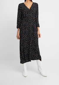 Robe noire à manches longues avec un col en V, taille cintrée, et imprimé floral coloré. Associée à des bottines blanches montant à la cheville. Tissu lisse, longueur midi.