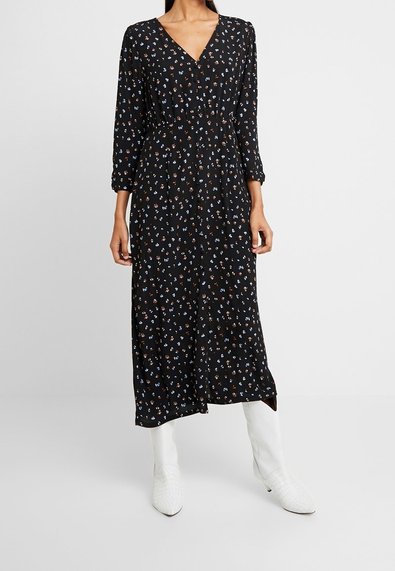 Robe noire à manches longues avec un col en V, taille cintrée, et imprimé floral coloré. Associée à des bottines blanches montant à la cheville. Tissu lisse, longueur midi.