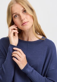 Pulover tricotat cu model cu nervuri de culoare albastru închis, cu decolteu larg și mâneci lungi. Textură netedă cu un detaliu subtil de cusătură pe față.
