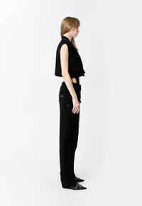 Chaleco negro corto con cuello, combinado con pantalones negros de talle alto. Los pantalones cuentan con bolsillos decorativos y zapatos de punta negra con tachuelas.