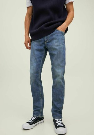 Jeans in denim azzurro chiaro con taglio dritto, dettagli sbiaditi e design classico a cinque tasche, abbinati a sneaker nere e a un gilet in maglia scuro.