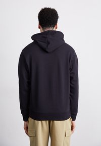 Sweatshirt negru cu glugă, cu o textură moale, având un buzunar tip cangur, manșete cu nervuri și o margine ușor scurtată.