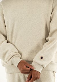 Pullover beige chiaro realizzato in tessuto morbido, caratterizzato da polsini e orlo a coste. Dettagli notevoli includono una toppa rotonda con logo sulla manica.