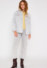 Giacca in denim grigia chiara con chiusura a bottoni e tasche, abbinata a jeans grigi chiari; il modello indossa una maglietta gialla e stivali con tacco marroni.