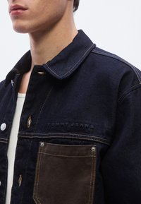 Σκούρο μπλε τζιν μπουφάν με μεγάλο γιακά, αντίθετη καφέ τσέπη, ραφές χαλκού και ανάγλυφο λογότυπο "TOMMY JEANS" στο στήθος.