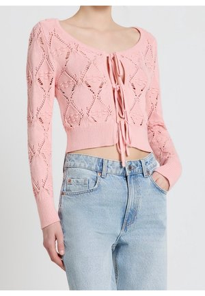 Femme portant un crop top en maille rose à manches longues avec des liens de fermeture, associé à un jean bleu délavé taille haute, main dans la poche.