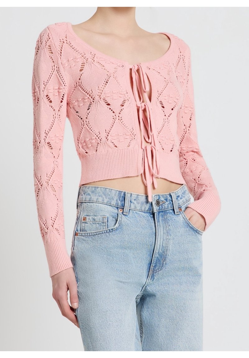 Femme portant un crop top en maille rose à manches longues avec des liens de fermeture, associé à un jean bleu délavé taille haute, main dans la poche.